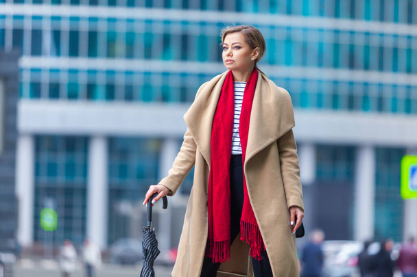Outdoors lifestyle fashion portrait of stunning brunette girl. Walking on the city street. Going shopping. Wearing stylish white fitted coat, red neckscarf, black umbrella cane. Business woman. - Fotó, kép