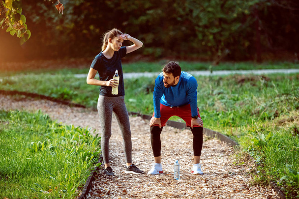 Fatigué couple sportif caucasien debout sur la piste dans les bois et se reposant de courir. Femme tenant bouteille avec rafraîchissement. Les deux sont en tenue de sport
. - Photo, image