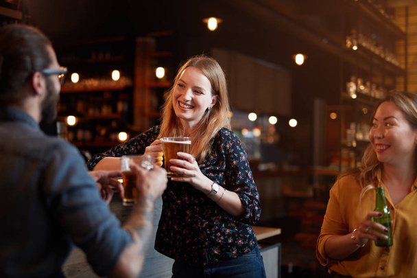 Усміхнена красива блондинка, спираючись на барну стійку, тримає пінту пива і розмовляє зі своїм другом. Нічне життя
. - Фото, зображення