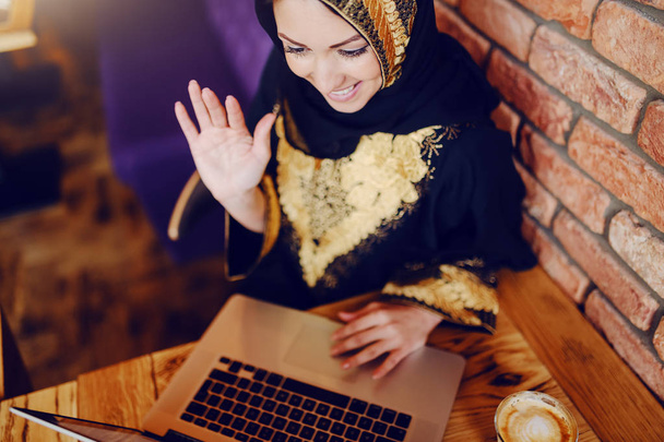 Widok z góry wspaniały uśmiechnięty muzułmanin kobieta ubrana w tradycyjny strój siedzi w kawiarni, mając połączenie wideo i machanie. Na drewnianym stole są laptop i kawa. - Zdjęcie, obraz
