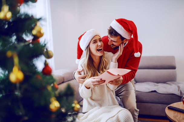 Stupita donna bionda caucasica carina seduta sul divano in soggiorno, abbracciando il suo ragazzo e ricevendo un regalo. Entrambi hanno cappelli da Babbo Natale in testa. In primo piano c'è l'albero di Natale. Soggiorno interno
. - Foto, immagini
