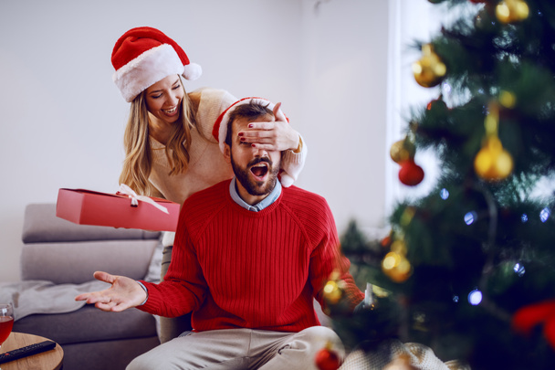 Jolie femme caucasienne souriante tenant cadeau et couvrant les yeux de son petit ami. Tous deux coiffés de chapeaux de Père Noël. Au premier plan se trouve l'arbre de Noël. Salon intérieur
. - Photo, image