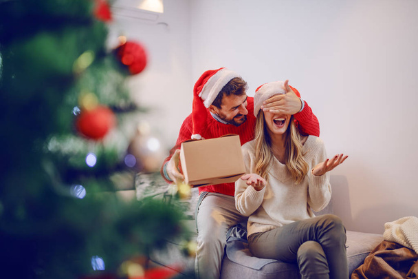 Smiling handsome bearded caucasian man covering his girlfriend's eyes and hiding gift behind back. Woman sitting on sofa. Both having santa hats on heads. Living room interior. - Zdjęcie, obraz