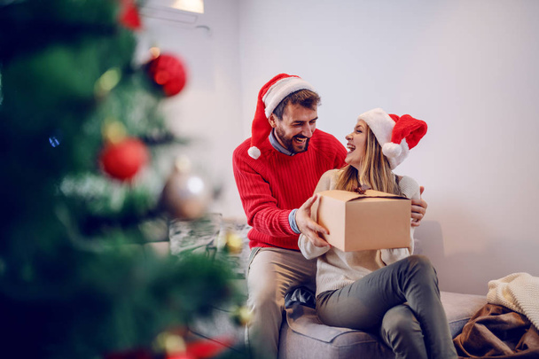 Amazed cute Caucasian blonde woman sitting on sofa in living room and receiving gift from her boyfriend. Both having santa hats on heads. In foreground is christmas tree. Living room interior. - Photo, Image