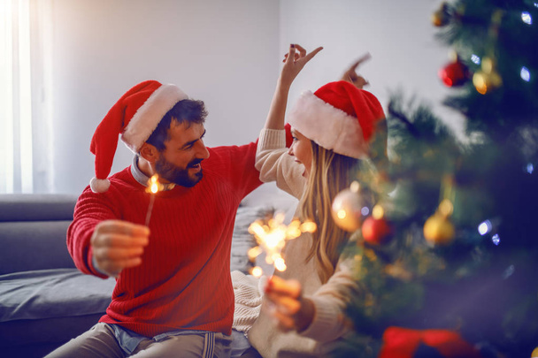 Szczęśliwy przystojny kaukaski para z Santa kapelusze na głowach siedzi na kanapie w salonie, świętując Nowy Rok i trzymając ognie. Na pierwszym planie jest choinka. - Zdjęcie, obraz