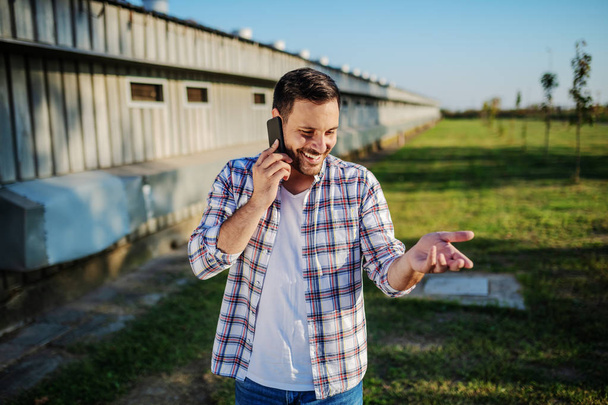 Ekose gömlekli ve kotlu yakışıklı beyaz çiftçi dışarıda duruyor ve telefonda konuşuyor. Arka planda ahır ve ağaçlar var. - Fotoğraf, Görsel