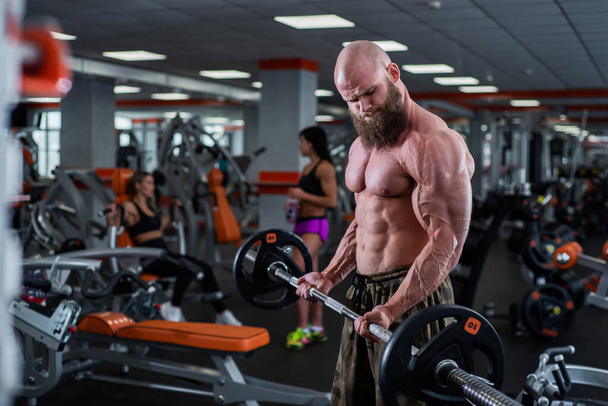 Bald man with a beard in the gym. Muscular bodybuilder guy doing barbell exercises. Strong man with a naked torso. The young athlete is preparing for weightlifting competitions. - Fotó, kép