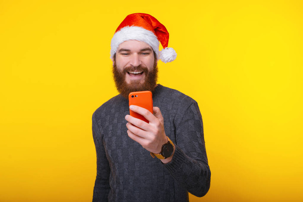 Uśmiechnięty młody brodaty mężczyzna w kapeluszu Świętego Mikołaja i z nowym telefonem komórkowym - Zdjęcie, obraz