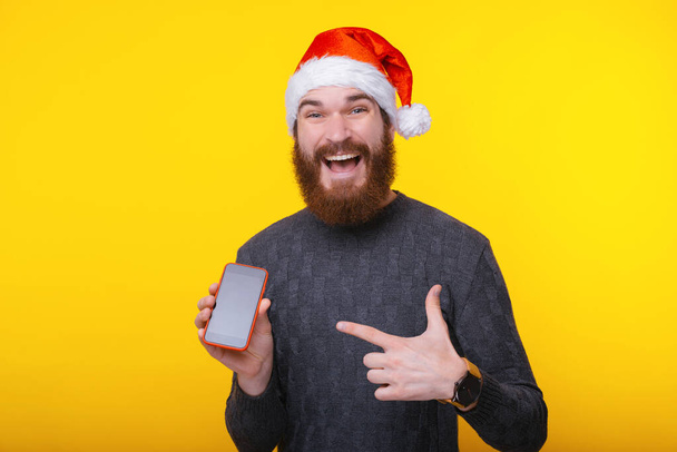 Happy bearded man wearing santa claus hat and pointing at smartphone screen - Foto, Imagen
