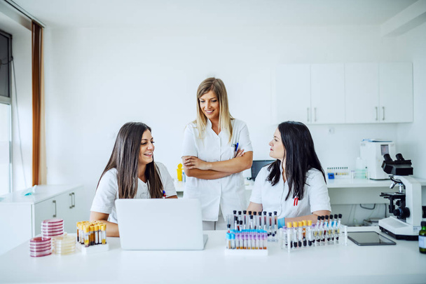 drei attraktive lächelnde kaukasische Laborassistentinnen in sterilen Uniformen, die im Labor sitzen und stehen, während sie an der Forschung arbeiten. Auf dem Labortisch liegen Reagenzgläser, Laptop, Petrischalen und Mikroskop. - Foto, Bild