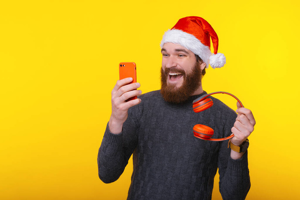 Excited man with beard wearing Santa Claus hat and looking at smartphone and holding red wireless headphones - Photo, Image