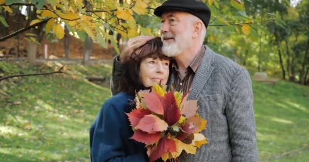 glückliche Eheleute, die sich unter einem Ast umarmen, den Herbst genießen, in einem gemütlichen Park zwischen den Bäumen - Zeitlupe. Frau hält einen Strauß Blätter in der Hand. - Filmmaterial, Video
