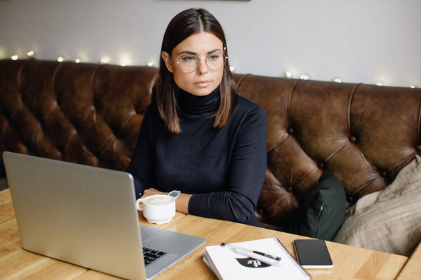 Young business woman sitting at table and taking notes in notebook.On table is laptop, smartphone and cup of coffee. Blogger - Zdjęcie, obraz