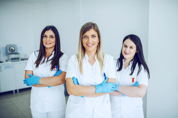 Trois beaux assistants de laboratoire caucasiens en uniformes blancs et avec des gants en caoutchouc debout dans le laboratoire avec les bras croisés
. - Photo, image