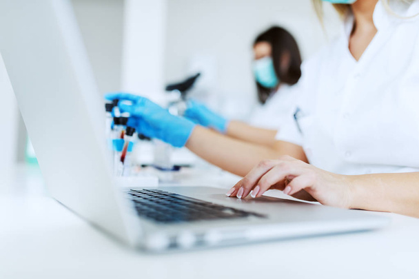 Close up of female lab assistant in white uniform sitting in lab and using laptop for data entry. In background is her colleague working. - Photo, Image