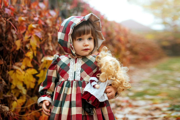 Little pretty girl in vintage dress with a porcelain doll in the autumn park - Photo, Image