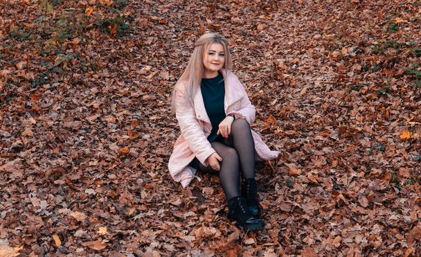 A young pleasant woman in casual clothes with long hair is sitting in the autumn forest. - Photo, Image