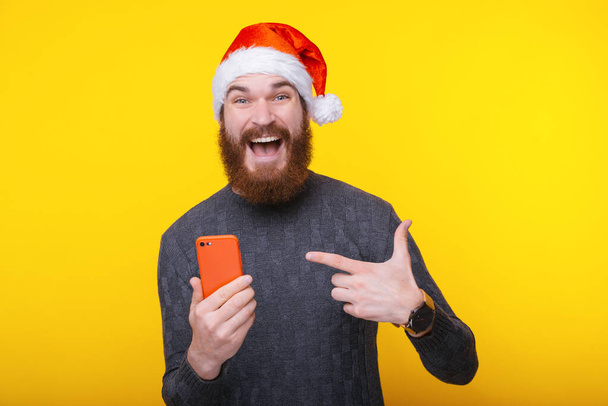 Amazed bearded hipster man in santa claus hat pointing red smartphone - Foto, Imagen