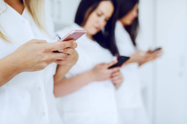 Група жінок-лаборантів у білій формі використовує смартфони на паузі, стоячи в лабораторії. Вибірковий фокус на руці зі смартфоном
. - Фото, зображення