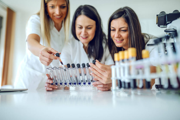 drei hingebungsvoll lächelnde kaukasische Laborassistentinnen, die Reagenzgläser mit Blutproben halten und betrachten. Laborinnenraum. Selektiver Fokus auf Ständer mit Reagenzgläsern. - Foto, Bild