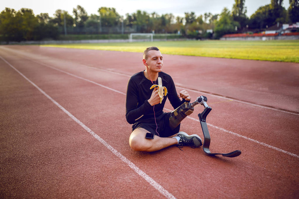 schöner kaukasischer sportlich behinderter junger Mann in Sportbekleidung und mit künstlichem Bein, der auf der Rennbahn sitzt, Musik hört und Bananen isst. - Foto, Bild