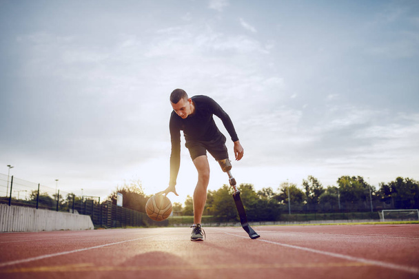 Πλήρες μήκος του αθλητισμού καυκάσιος ανάπηρος άνθρωπος στα αθλητικά και τεχνητό πόδι dribble την μπάλα, ενώ στέκεται σε πίστα. - Φωτογραφία, εικόνα
