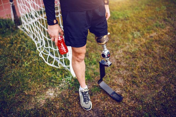 Knappe sportieve gehandicapte man in sportkleding en met kunstbeen terwijl hij op het voetbalveld staat en een verfrissing vasthoudt. - Foto, afbeelding