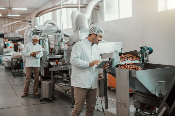 Young Caucasian supervisor evaluating quality of food in food plant while holding tablet. Man is dressed in white uniform and having hair net. - Photo, Image