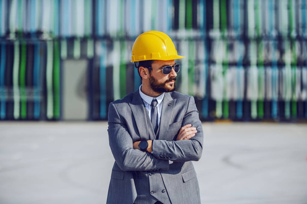 Empresário barbudo caucasiano sério elegante em terno, com óculos de sol e capacete na cabeça em pé no local de construção com os braços cruzados e supervisionando as obras
. - Foto, Imagem