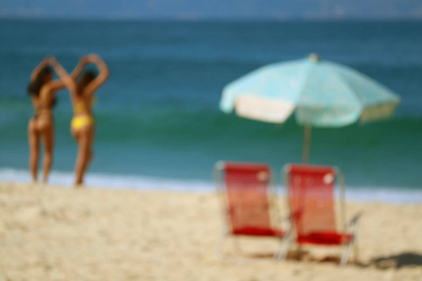 Blurred image of a pair of red beach chairs and light blue parasol on the sandy beach against vivid blue ocean - Photo, Image