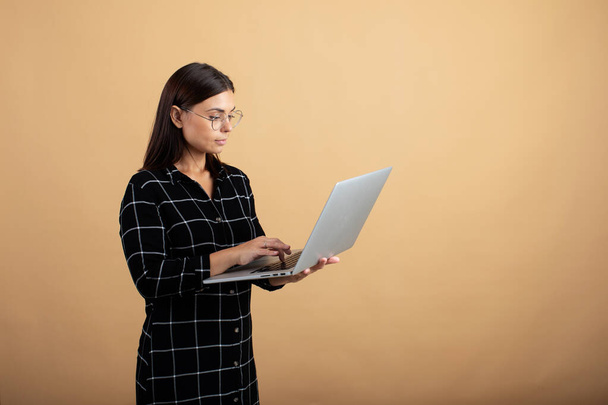A young woman in a plaid dress stands on an orange background with a laptop - Foto, Bild