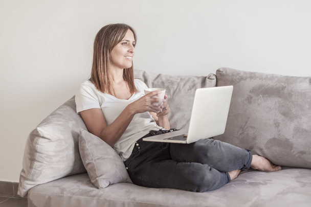 Adult woman sitting on a sofa while using laptop and holding cup of tea - Foto, immagini
