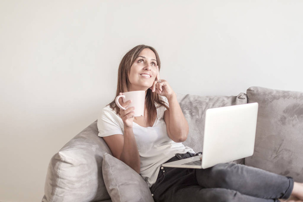 Kaunis aikuinen nainen rentouttava sohvalla tilalla kuppi kahvia käytettäessä kannettavaa tietokonetta
 - Valokuva, kuva