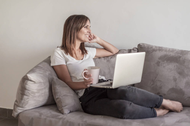 Beautiful adult woman relaxing on the sofa holding a cup of coffee while using laptop - Фото, изображение