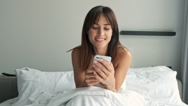 Happy beautiful young brunette woman using smartphone while sitting in bed in the morning - Video, Çekim