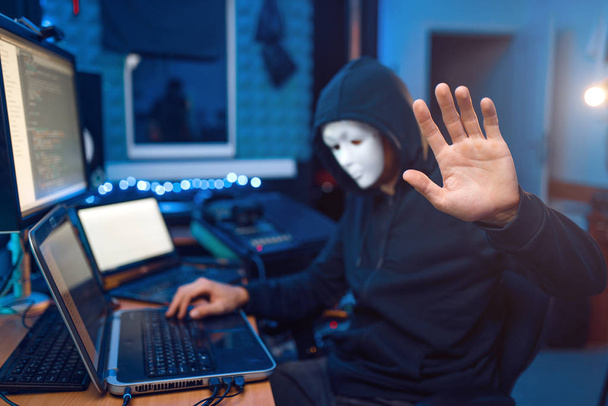 Хакер в маске и капюшоне сидит на своем рабочем месте с ноутбуком и компьютером, паролем или взломом учетной записи. Интернет-шпион, нелегальный образ жизни, рискованная работа, сетевой преступник
 - Фото, изображение