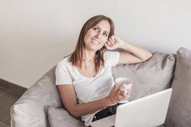 Beautiful adult woman relaxing on the sofa holding a cup of coffee while using laptop - Fotografie, Obrázek
