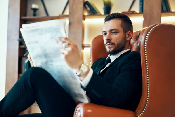 Уверенный молодой бизнесмен читает газету и последние новости, сидя в кресле
 - Фото, изображение