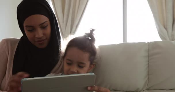 Front view of a young mixed race woman wearing hijab with her young daughter in the sitting room, sitting on a sofa and using a tablet computer - Video, Çekim