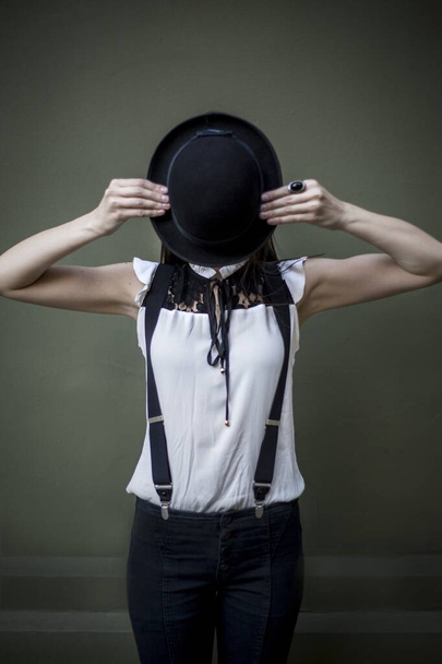 A vertical shot of a female in a white shirt and black trousers holding a black hat in front of her face - Фото, зображення