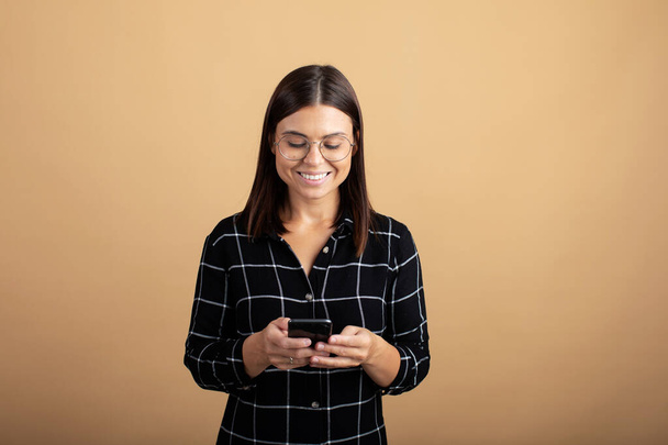 A young woman in a plaid dress stands on an orange background and plays with her phone - Φωτογραφία, εικόνα