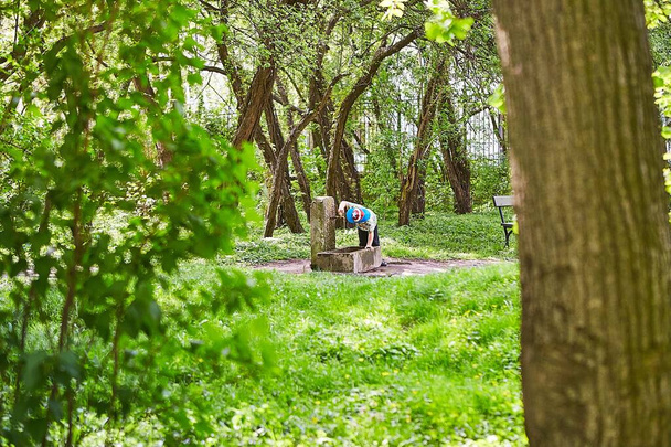 Person with a blue hat drinking water from a water fountain surrounded by green trees - 写真・画像