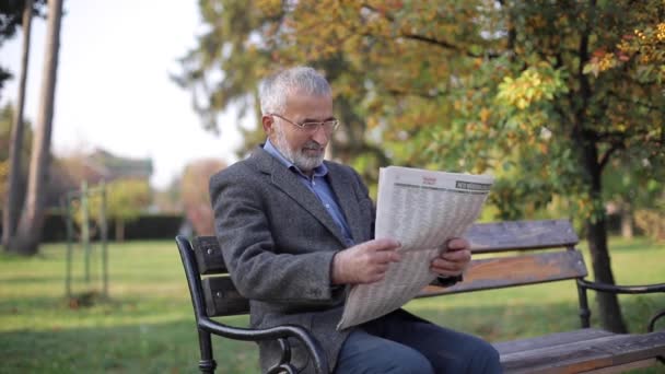 Bearded elderly man in glasses reading newspaper in the autumn park. Handsome gray-haired man sitting on the bench early in the morning - Filmagem, Vídeo