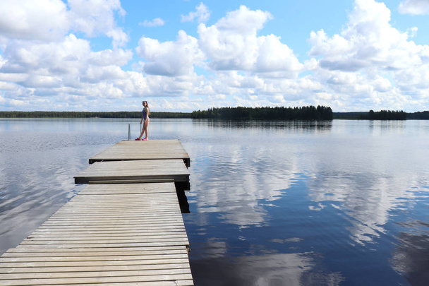 Adolescente (14) parada en un muelle en el lago Ranuanjrvi con reflejos del cielo y nubes en el agua en un soleado día de verano en Ranua, Laponia en el norte de Finlandia
.  - Foto, imagen