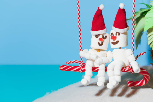 Two lovers of Marshmallow snowmen are sitting on a swing of candy. Christmas holidays on the beach. - Photo, Image
