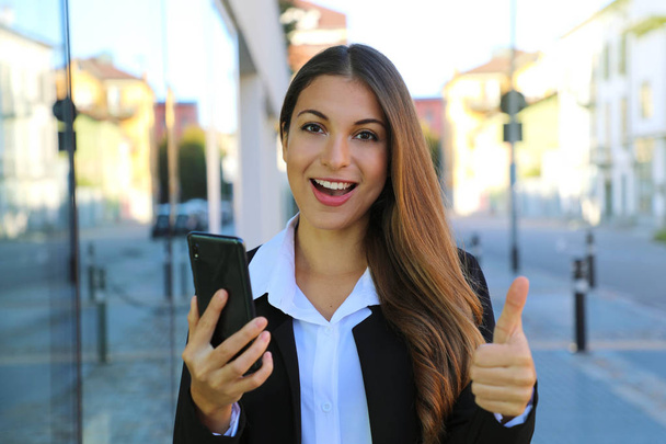 Pretty beautiful young business woman holding smart phone gives thumb up in the street. - Fotografie, Obrázek