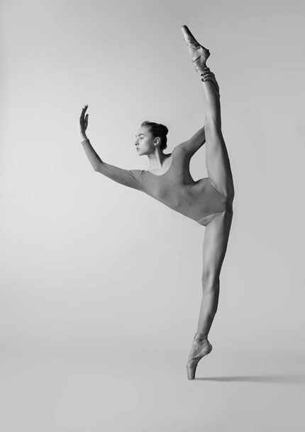 Balerina spárgázik, miközben szürke tornadresszben és pointe-ban áll. Fekete-fehér fénykép. - Fotó, kép