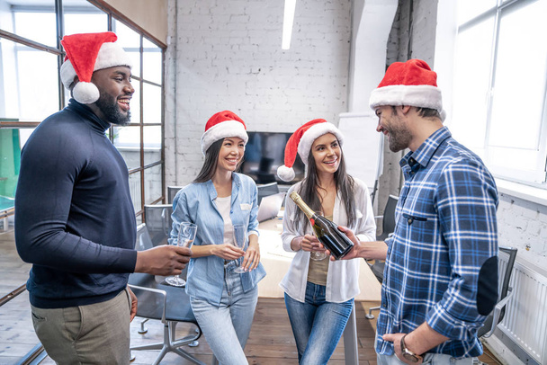 Mutlu Noeller ve mutlu yıllar 2020. Çok ırklı genç yaratıcı insanlar modern ofiste bayramı kutluyorlar. Bir grup genç iş adamı şampanya içiyor ve birlikte çalışarak eğleniyorlar.. - Fotoğraf, Görsel