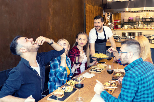 小さなレストランでおいしい料理を食べる親友のグループと幸せな所有者がそれらを提供しています-流行のパブで一緒に時間を楽しんでいる若者たちは赤ワインを飲んで、ウェイターが提供する試飲スナック - 写真・画像
