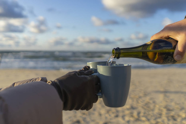 żeńskie dłonie w brązowych skórzanych rękawiczkach trzymają kubki nad morzem w słoneczny jesienny dzień nad morzem, nalewają szampana - Zdjęcie, obraz